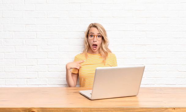 young blonde woman looking shocked and surprised with mouth wide open, pointing to self using a laptop - Foto, Imagem