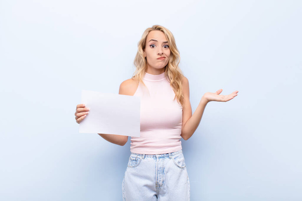 young blonde woman feeling puzzled and confused, doubting, weighting or choosing different options with funny expression holding a sheet of paper - Fotó, kép