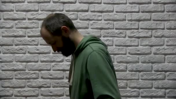 Ein Typ in grünem Sweatshirt mit Kapuze und Bart steht vor weißem Backsteinhintergrund, nimmt ein rotes, großes Tonbandgerät, legt es auf die Schulter, schaut sich um und geht hinterher - Filmmaterial, Video