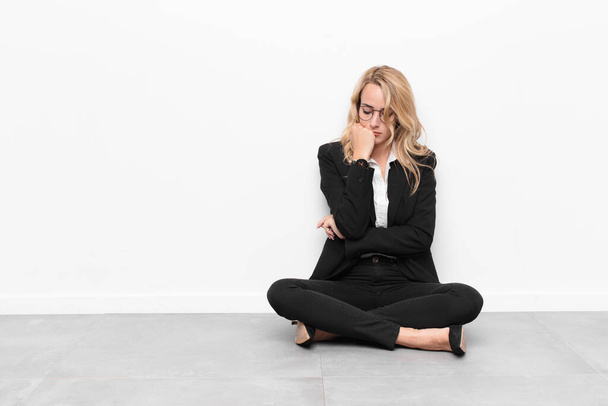 young blonde woman feeling serious, thoughtful and concerned, staring sideways with hand pressed against chin sitting on the floor - Foto, imagen