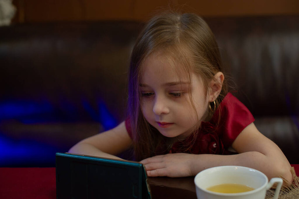 Mała dziewczynka w burgundowej sukience patrzy na telefon. Dziecko dziewczyna 4 lub 5 lat w kawiarni lub w domu oglądając coś ciekawego w smartfonie. Dziecko pije zieloną lub ziołową herbatę. Dzieciństwo - Zdjęcie, obraz