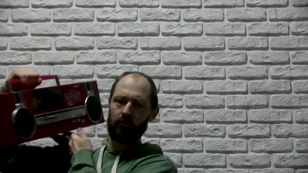 een man in een groene sweater met een capuchon en een baard staat tegen een achtergrond van witte bakstenen, pakt een rode oude grote tape recorder en zet het op zijn schouder, kijkt rond en gaat achter - Video