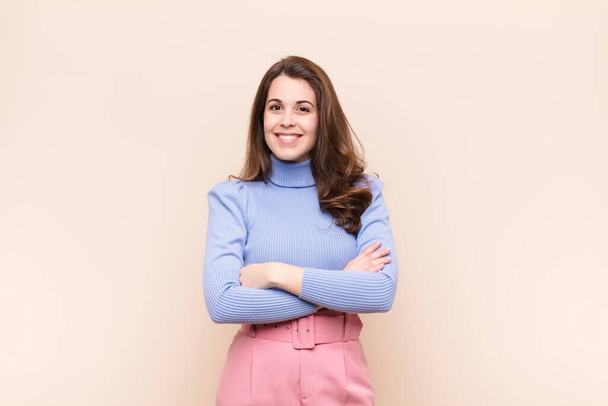 young pretty woman smiling to camera with crossed arms and a happy, confident, satisfied expression, lateral view against beige background - Foto, Imagen