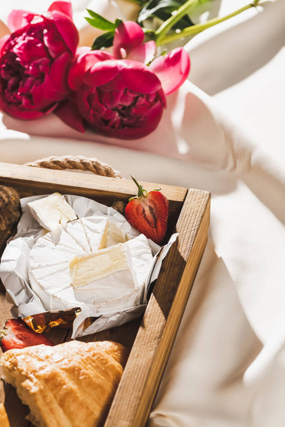 close up view of french breakfast with croissant, Camembert, strawberries on wooden tray on textured white cloth with peonies - Fotografie, Obrázek