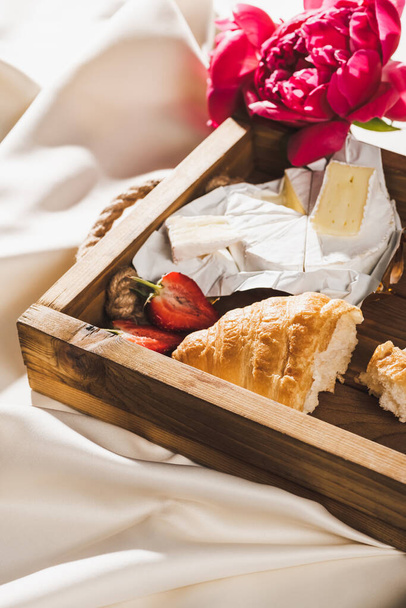 close up view of french breakfast with croissant, Camembert, strawberries on wooden tray on textured white cloth with peonies - Photo, Image