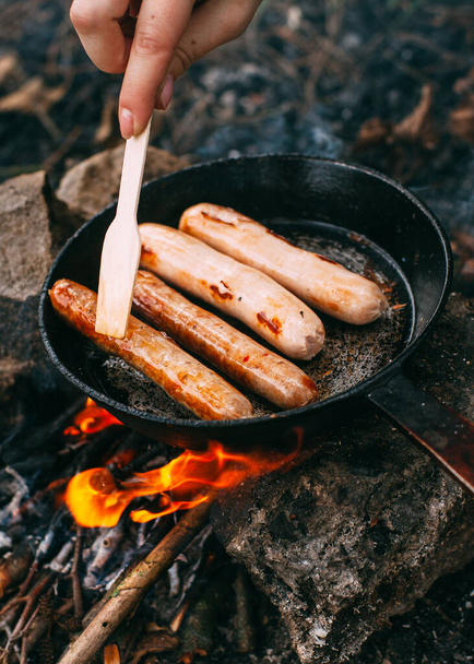 Roasting sausages in a frying pan over an open fire. Preparing food in nature. Lunch in the open air. Hand with a wooden shovel. Picnic in the forest. Food on fire - Photo, Image