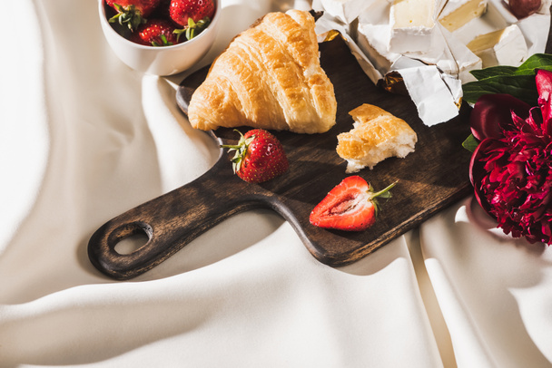 top view of french breakfast with strawberries, croissant, Camembert, peony on wooden cutting board on white tablecloth - Foto, Imagen