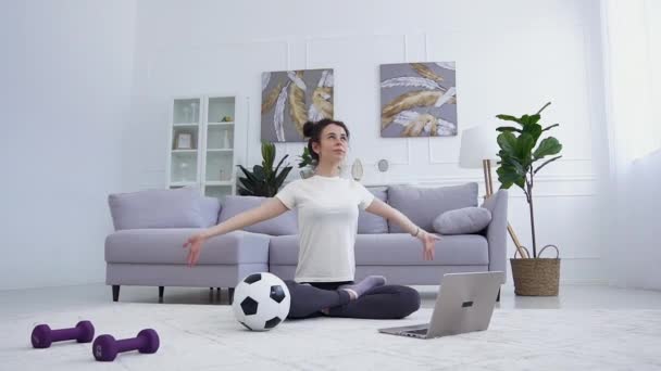 Front view of charming calm happy young brunette in sportwear which sitting on the floor at home in lotus pose and doing yoga exercises for breath - Materiaali, video