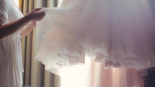 Střední pohled na evropskou tisíciletou nevěstu v elegantním bílém peignoiru, dotýkající se, vonící a pociťující luxusní velké svatební šaty visící poblíž záclon místnosti pod jasným osvětlením. - Záběry, video