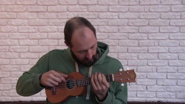 portret jednego młodego człowieka, który siedzi przed kamerą i gra na ukulele, pokazuje różne emocje, jest zaskoczony, na tle białej ściany - Materiał filmowy, wideo