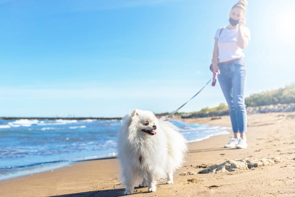 Chica con máscara protectora paseando con perro en la playa. Perro pomerano blanco
. - Foto, imagen