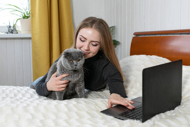 The girl lies on the bed with a laptop. Hugs a gray cat and looks into the laptop. Shopping for pets. Onlan shopping. Online training. Distance learning. We are sitting at home. Purebred cat - Фото, изображение