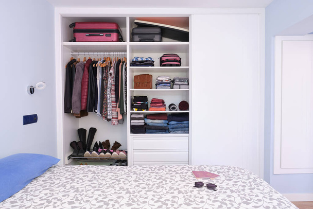 Modern hálószoba belső tér nyitott szekrény tele színes ruhák és bőröndök. Útlevelek és napszemüveg az ágyon. - Fotó, kép