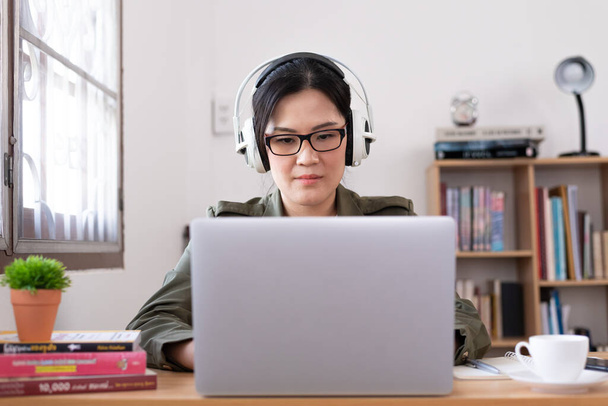 Современная молодая азиатка работает из дома и слушает музыку в белых наушниках
. - Фото, изображение