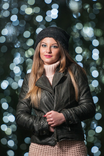 Fashionable lady wearing cap and black jacket outdoor in xmas scenery with blue lights in background. Portrait of young beautiful woman with long fair hair posing smiling in winter style. - 写真・画像