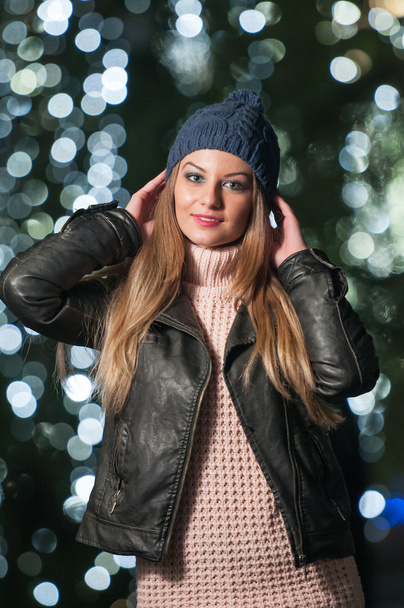 Fashionable lady wearing cap and black jacket outdoor in xmas scenery with blue lights in background. Portrait of young beautiful woman with long fair hair posing smiling in winter style. - Foto, immagini