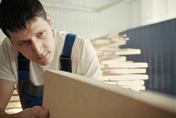 Un giovane falegname sceglie una tavola di legno. Ispezione e controllo qualità del legno. Lavoratore nella falegnameria
. - Foto, immagini
