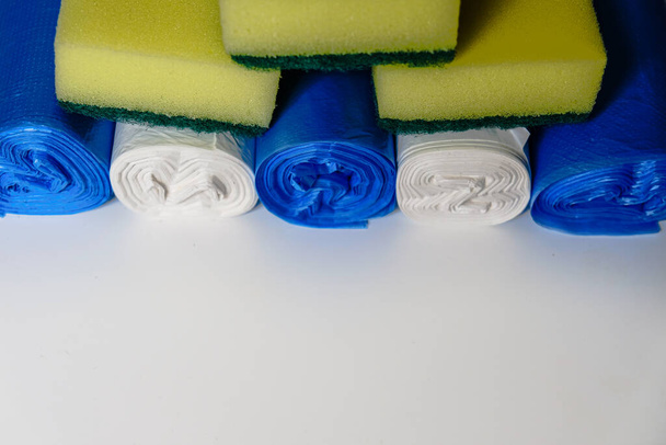 Schoonmaakfaciliteiten tijdens isolatie van het coronavirus. Gele schotel wassen sponzen en kleurrijke plastic vuilniszakken. Dagelijkse huisvrouw keukenapparatuur en essentiële gereedschappen gestapeld op een wit oppervlak. - Foto, afbeelding