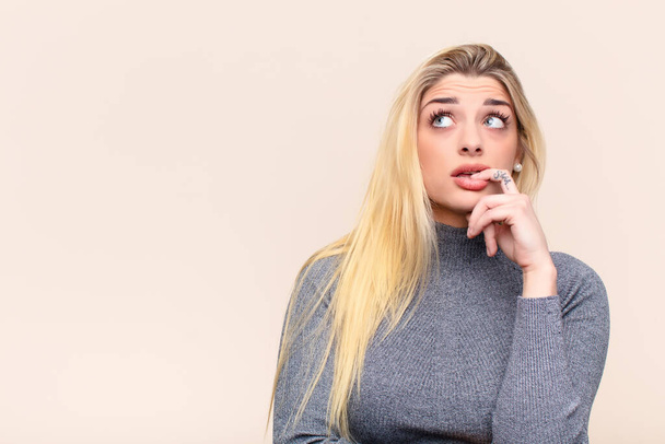 young pretty blonde woman with surprised, nervous, worried or frightened look, looking to the side towards copy space against flat wall - Photo, Image