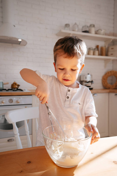 Parlak bir mutfağın arka planında bir çocuk cam bir tabakta yemek pişirmek için hamur yoğurur. Bir çocuk annesine yemek pişirmesinde yardım eder.. - Fotoğraf, Görsel