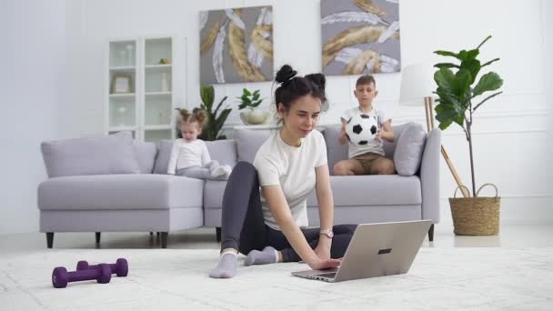 Family idyll where good-looking calm different ages children sitting on the sofa while their sportive smiling cute mother doing fitness stretching exercises on the carpet near laptop - Filmati, video
