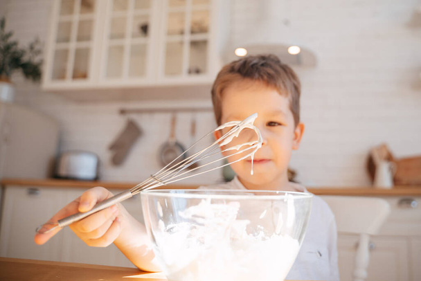 Un niño en el fondo de una cocina brillante amasa una masa para hornear en un plato de vidrio.Desenfoque de cerca en un batidor con masa goteante
. - Foto, Imagen