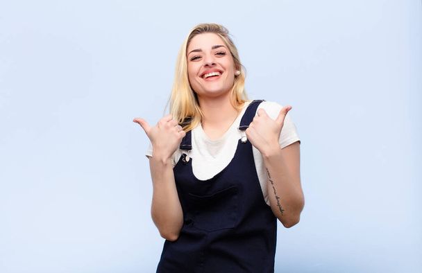 young pretty blonde woman smiling joyfully and looking happy, feeling carefree and positive with both thumbs up against flat wall - Foto, Imagen