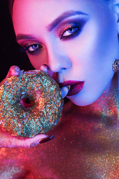 Portrait of Beautiful Woman with Sparkles on her Face and donut in hand. Girl with Art Make-Up in Color Light. - Foto, Bild