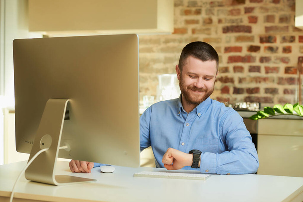 A happy man with a beard looking at his wristwatch during an online video briefing with his colleagues at home. A guy with a smile working remotely on a desktop computer in a private apartment. - Photo, Image