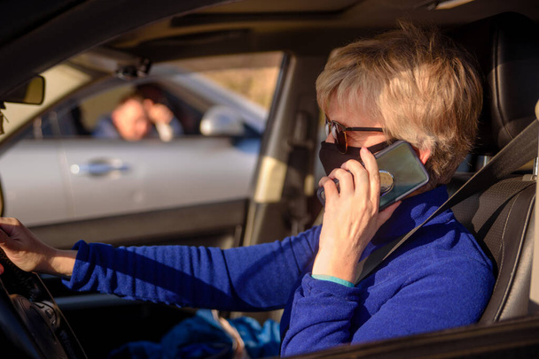 La conductrice en masque noir et lunettes de soleil conduit une voiture et parle au téléphone. Conduite à droite. Concept de lutte contre le coronavirus
. - Photo, image