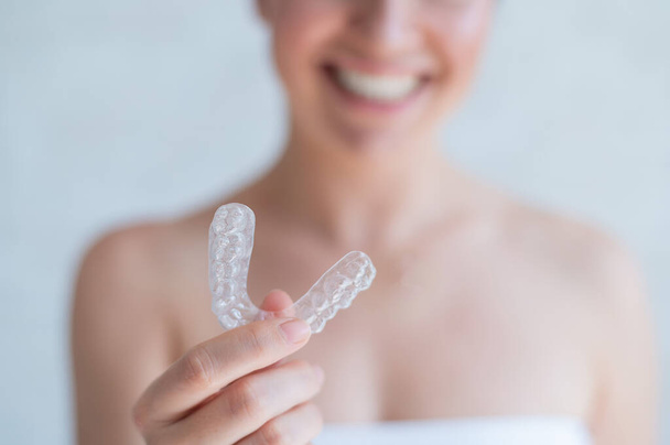 Close-up of orthodontic silicone transparent teeth aligner in female hands. Blurred unrecognizable woman in a white towel holds a removable night retainer. Brace for teeth whitening. - Фото, изображение