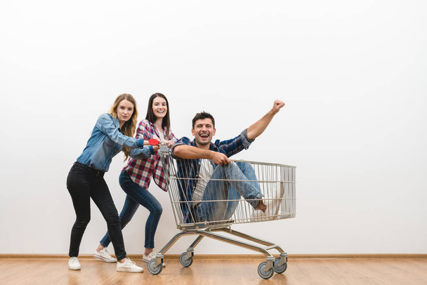 The two women and a man fun with a shopping cart on a white wall background - Photo, Image