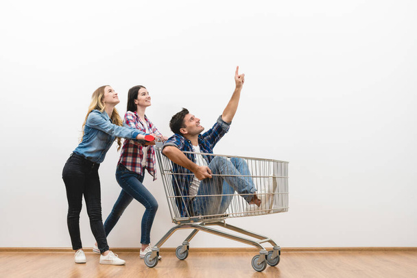 Die drei glücklichen Menschen amüsieren sich mit einem Einkaufswagen auf weißem Wandhintergrund - Foto, Bild