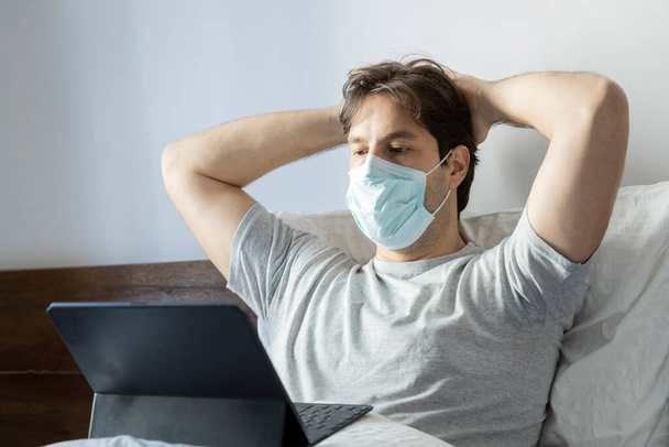 Hombre trabajando en la cama, haciendo oficina en casa porque está enfermo. usando una máscara sobre la boca y la nariz
. - Foto, Imagen