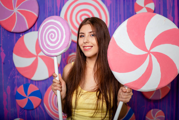 Egy gyönyörű, fiatal nő egy nagy játékcukorkával a kezében, egy absztrakt lila-kék háttéren áll, édességekkel és nyalókákkal.. - Fotó, kép