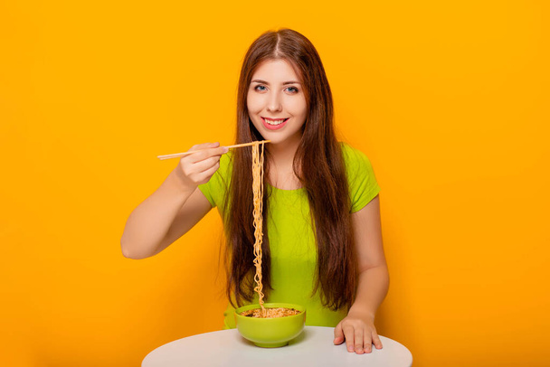 Привлекательная молодая женщина ест лапшу с палочками для еды из зеленой миски, сидя за белым столом на желтом фоне
. - Фото, изображение