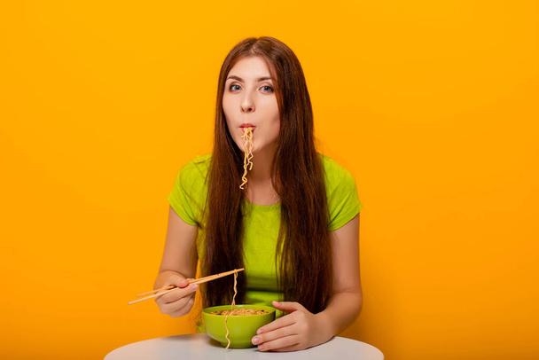 Attractive, young woman eating instant noodles with chopsticks from a green bowl, sitting at a white table on a yellow background. - Foto, immagini