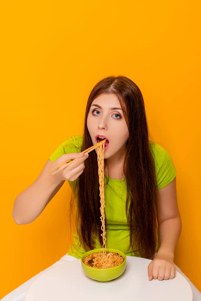 黄色の背景に白いテーブルに座って、緑のボウルから箸でインスタントラーメンを食べる魅力的な若い女性. - 写真・画像