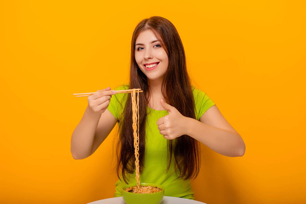 Aantrekkelijke, jonge vrouw die instant noedels eet met eetstokjes uit een groene kom, zittend aan een witte tafel op een gele achtergrond. - Foto, afbeelding