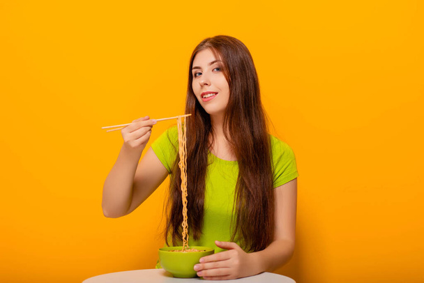 Vonzó, fiatal nő eszik instant tészta evőpálcikával egy zöld tál, ül egy fehér asztal sárga háttér. - Fotó, kép