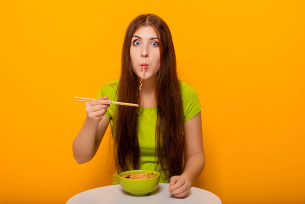 Ελκυστική, νεαρή γυναίκα τρώει στιγμιαία νουντλς με ξυλάκια από ένα πράσινο μπολ, κάθεται σε ένα λευκό τραπέζι σε κίτρινο φόντο. - Φωτογραφία, εικόνα