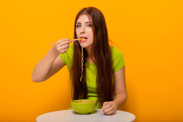 Привлекательная молодая женщина ест лапшу с палочками для еды из зеленой миски, сидя за белым столом на желтом фоне
. - Фото, изображение