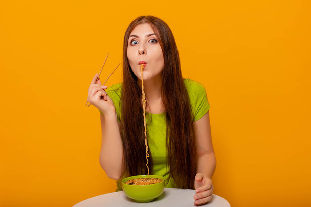 Mulher jovem e atraente comendo macarrão instantâneo com pauzinhos de uma tigela verde, sentada em uma mesa branca em um fundo amarelo
. - Foto, Imagem