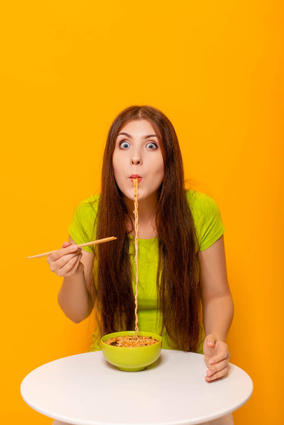Attractive, young woman eating instant noodles with chopsticks from a green bowl, sitting at a white table on a yellow background. - Foto, afbeelding