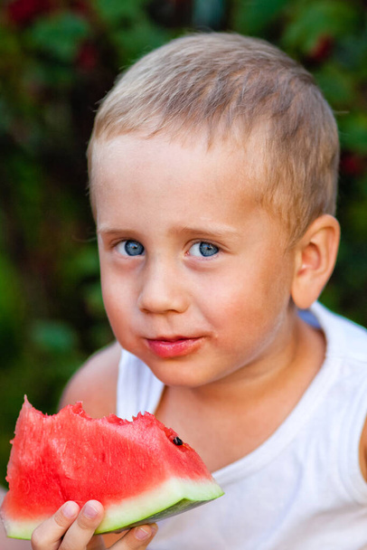 Chlapeček sní kousek zralého červeného melounu. Šťastné dětství v létě na dvorku. Kavkazský chlapec má venkovní piknik na travnatém trávníku - Fotografie, Obrázek