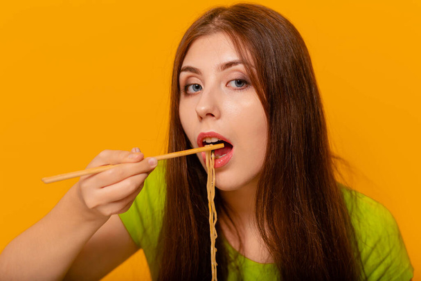 Woman eating instant noodles with chinese chopsticks. Studio photo on a yellow background. - 写真・画像