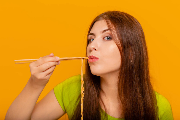 Woman eating instant noodles with chinese chopsticks. Studio photo on a yellow background. - Foto, Imagen