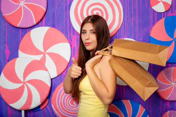 Young, attractive woman with paper bags in her hands on a background of a purple wall made of sweets and lollipops. - Foto, Bild