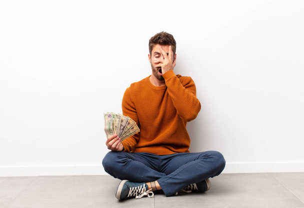 young handsome man looking shocked, scared or terrified, covering face with hand and peeking between fingers holding dollar banknotes - Фото, изображение