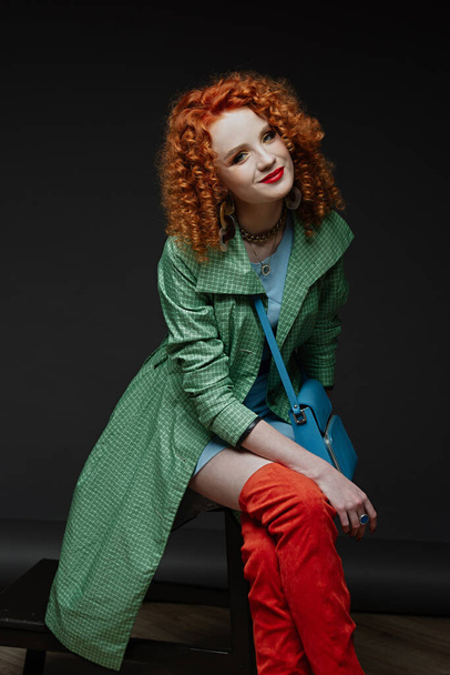the girl smiles, curly hair, the red-haired girl sits on a chair, crosses her legs, portrait in a photo studio, bright clothes, orange boots, magnificent hair, a green alash - 写真・画像